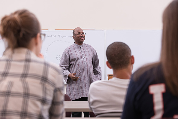 A smiling male professor teaching a group of students in front of a whiteboard during a religious studies class