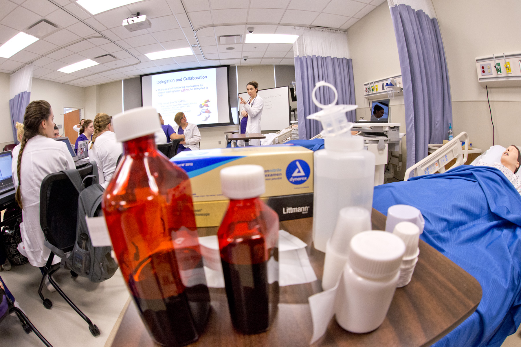 Students in white coats listen to a female professor in front of a projector in a nursing simulation lab surrounded by medical equipment and prescription bottles