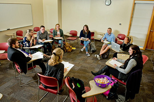 A female professor sitting in a round classroom configuration with several students discussing public relations