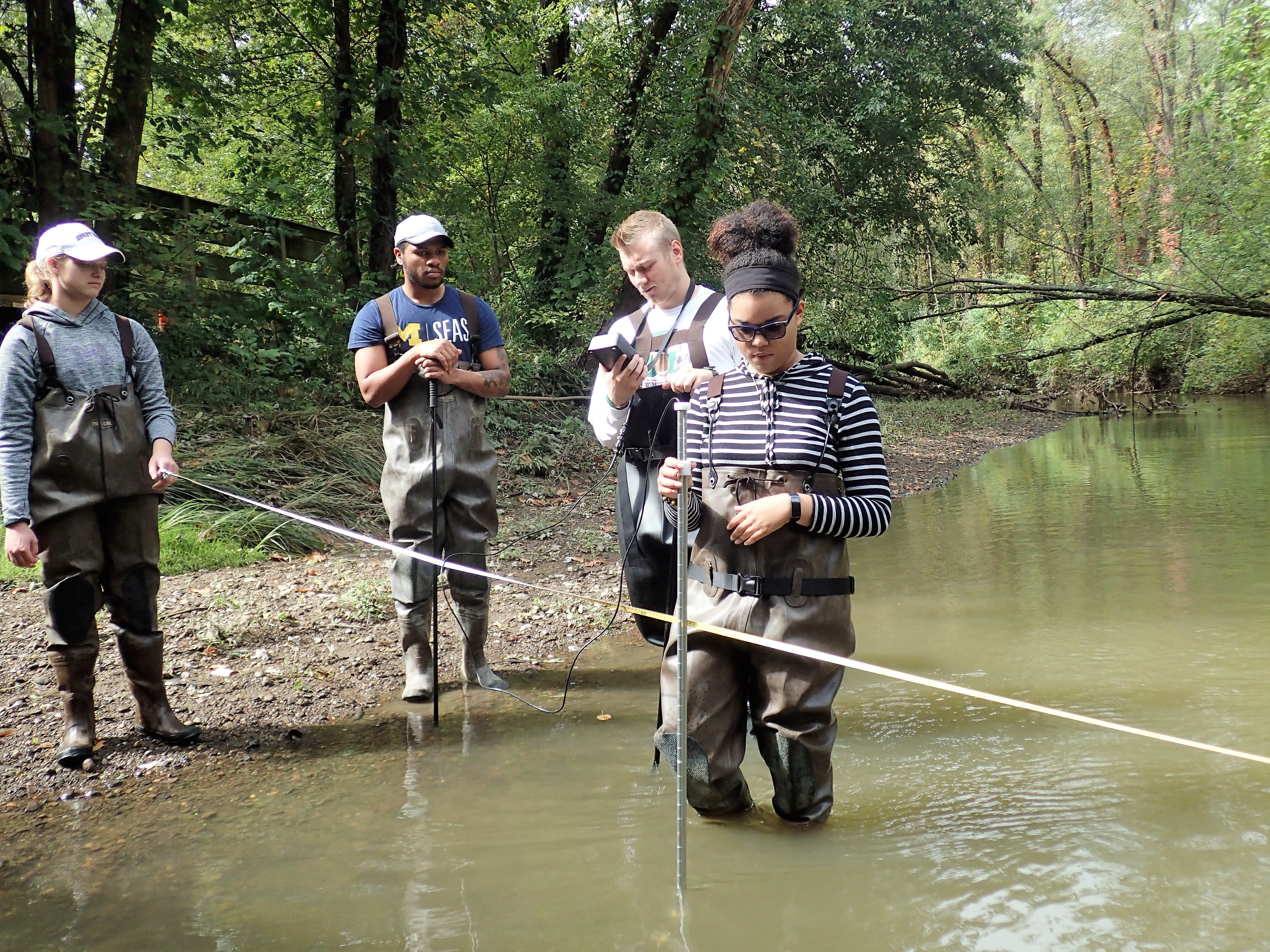 Four environmental science students wade in the water with a rope and measurement tools to evaluate river flow