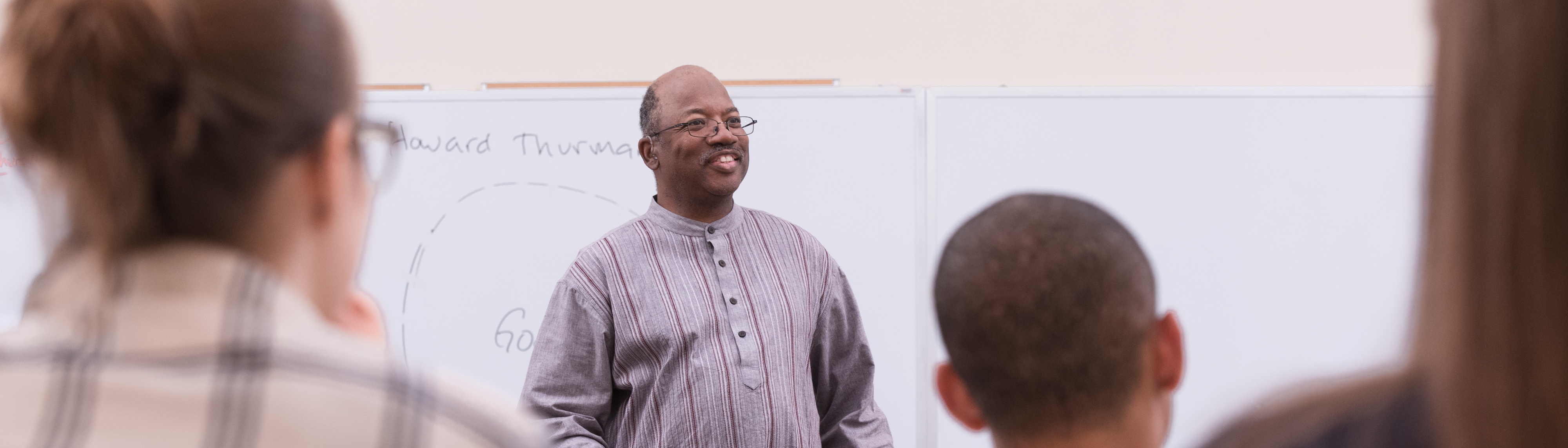 A smiling male professor teaching a group of students in front of a whiteboard during a religious studies class