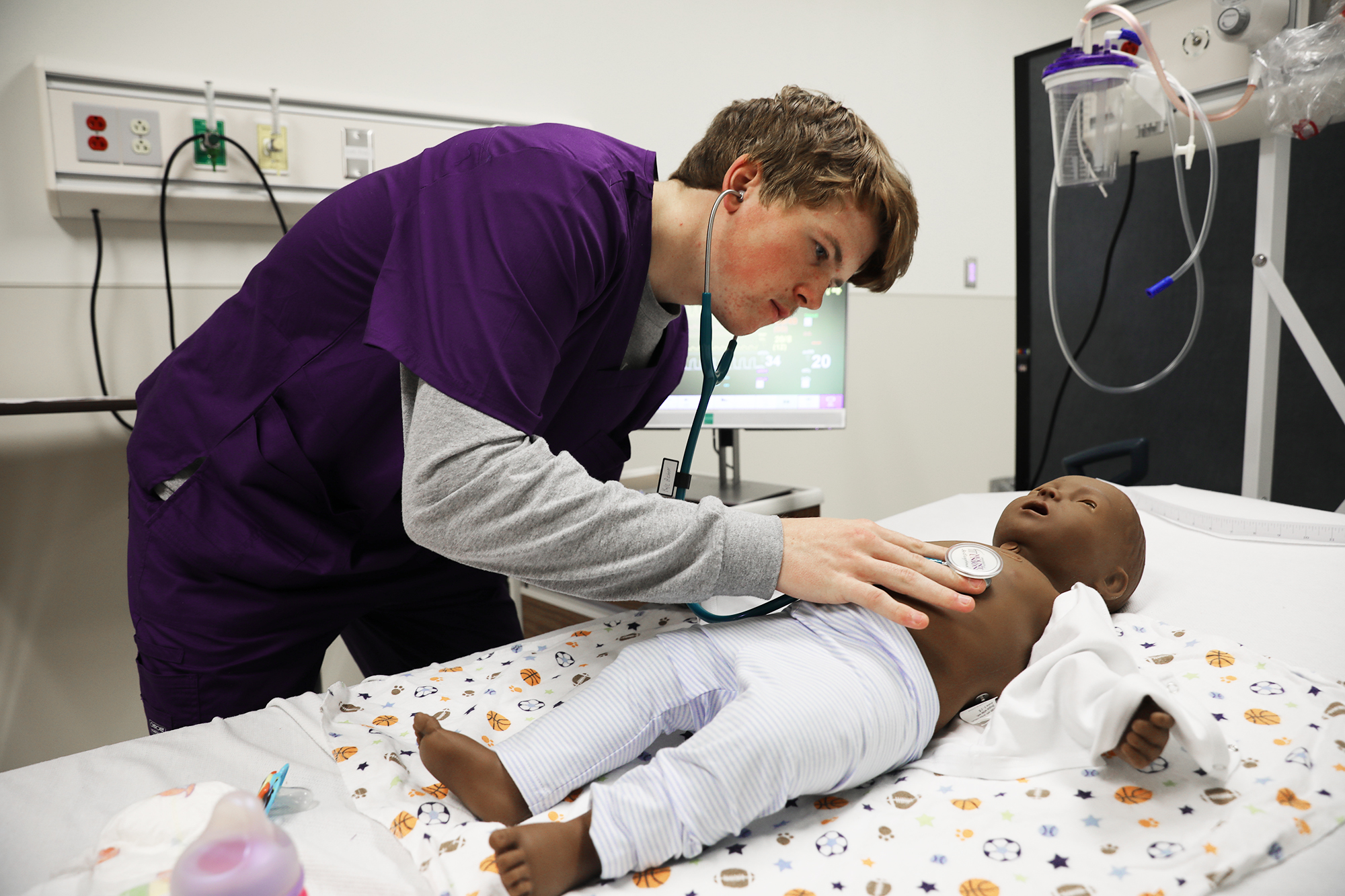A student uses a stethoscope in a nursing lab to listen for to a simulation infant's heartbeat
