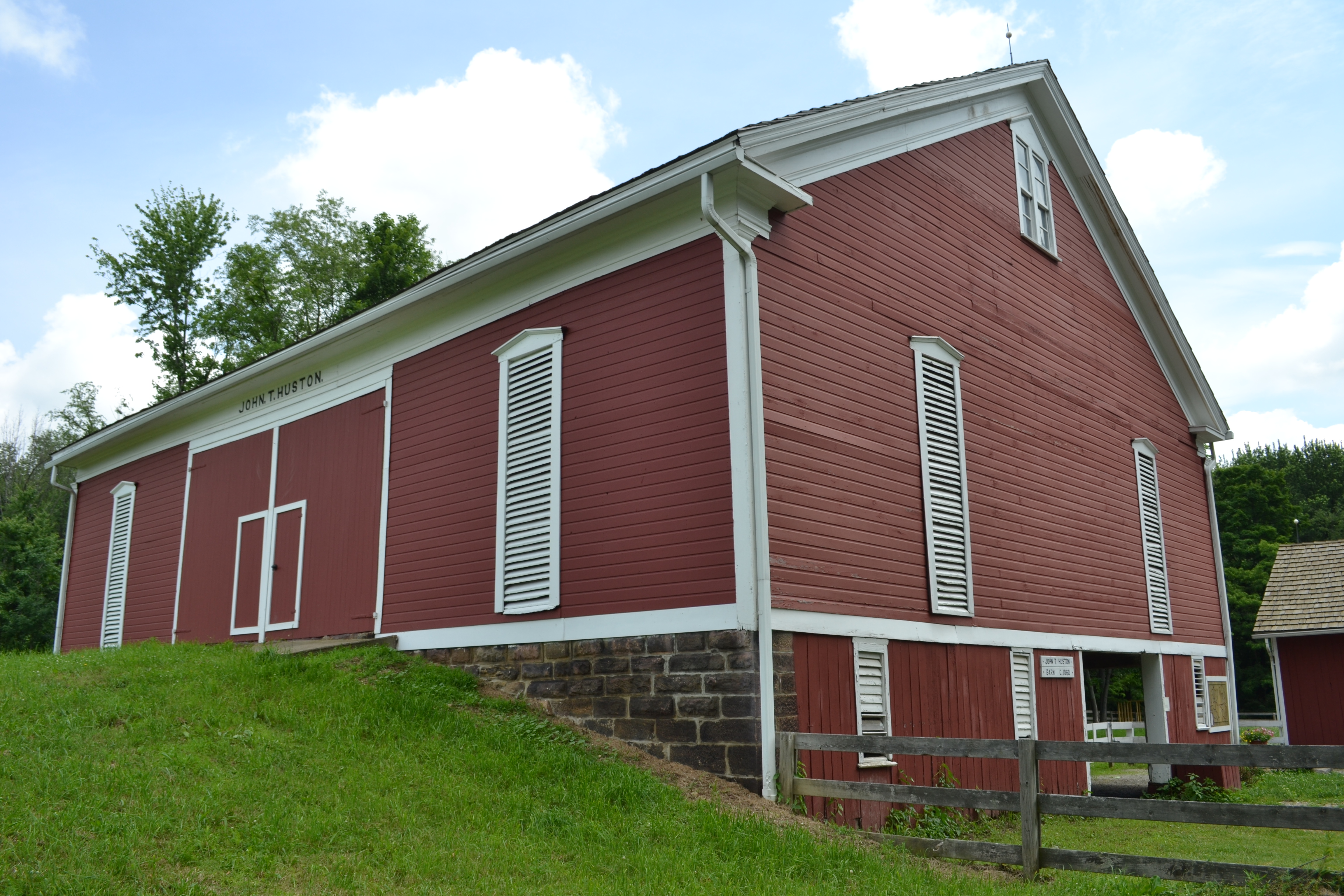 A large, red barn with white trim and louvered windows. The barn is built on a stone foundation. There's a wooden fence in front of the barn, and green grass surrounds it.
