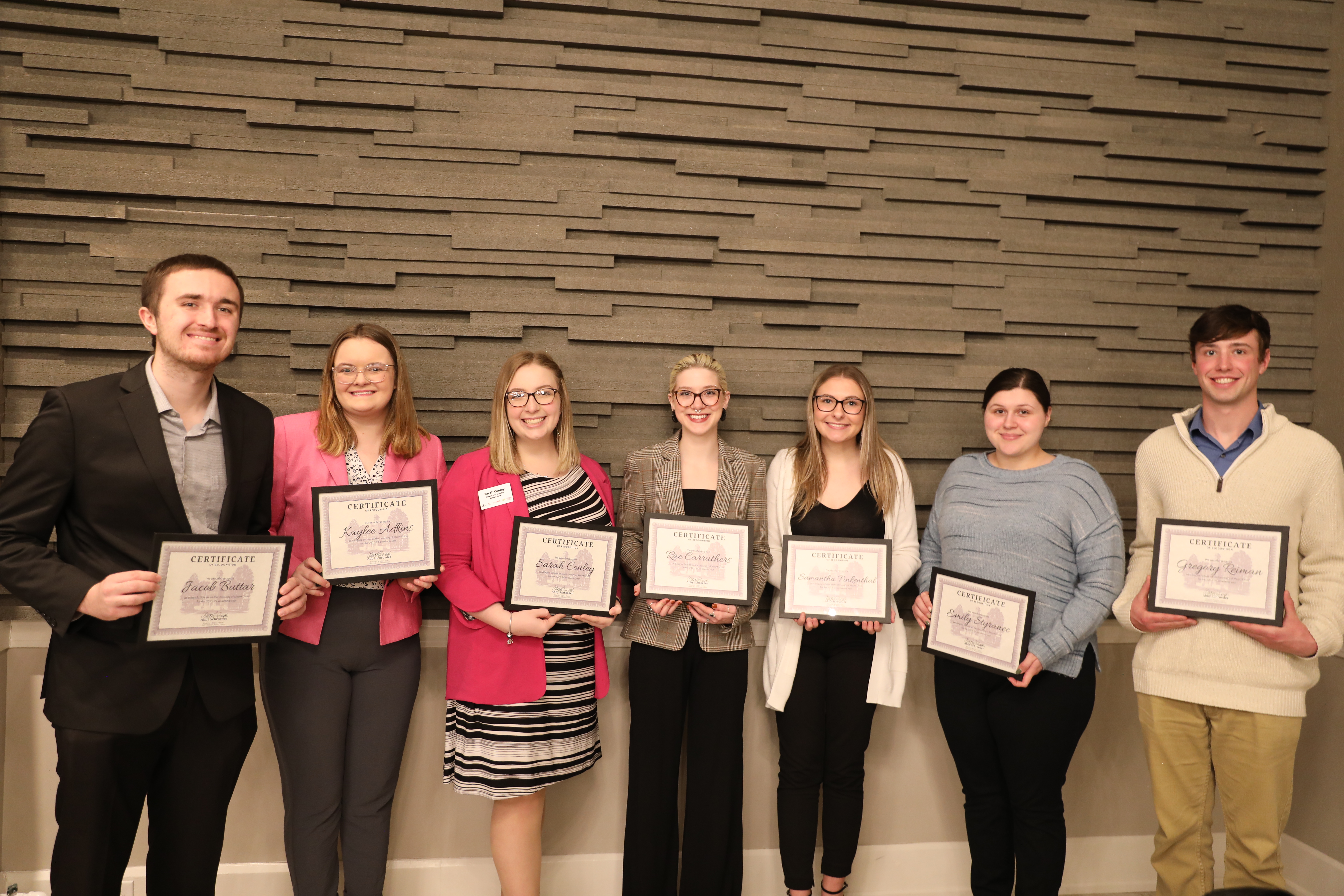 Seven students stand holding Regula Scholar certificates bearing their names while smiling in business casual attire