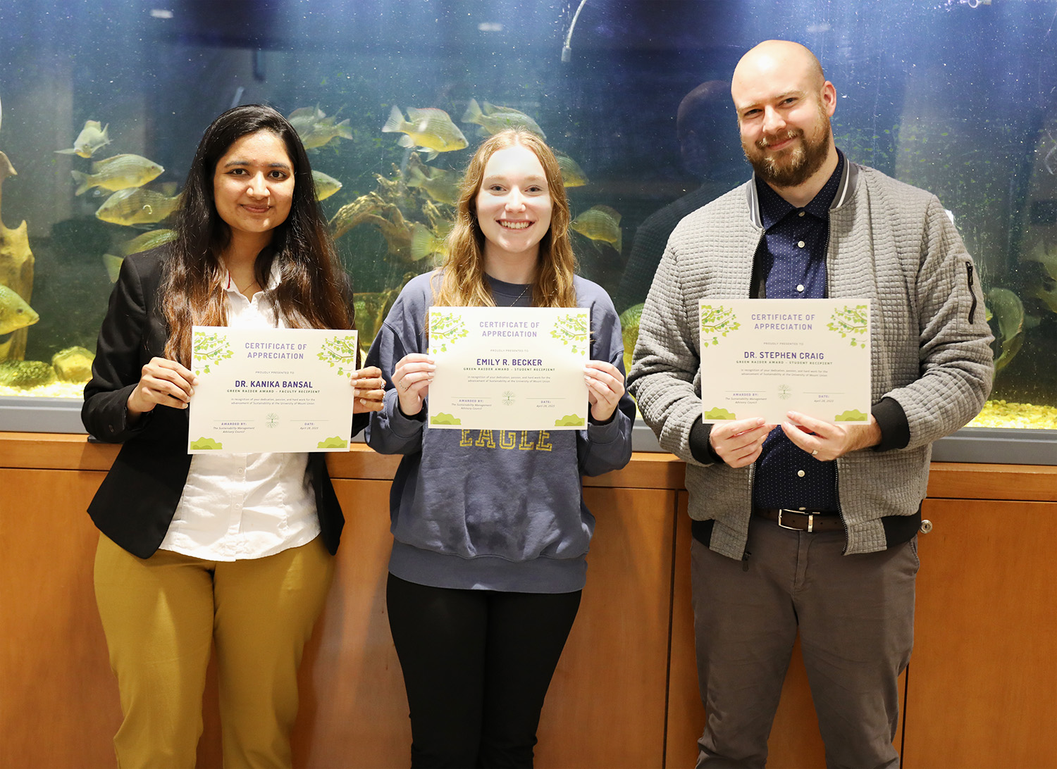From left to right: Dr. Kanika Bansal , Emily C. Becker, and Dr. Stephen Craig stand smiling while holding their Green Raider Award Certificates in front of an aquarium in the Bracy Hall science building