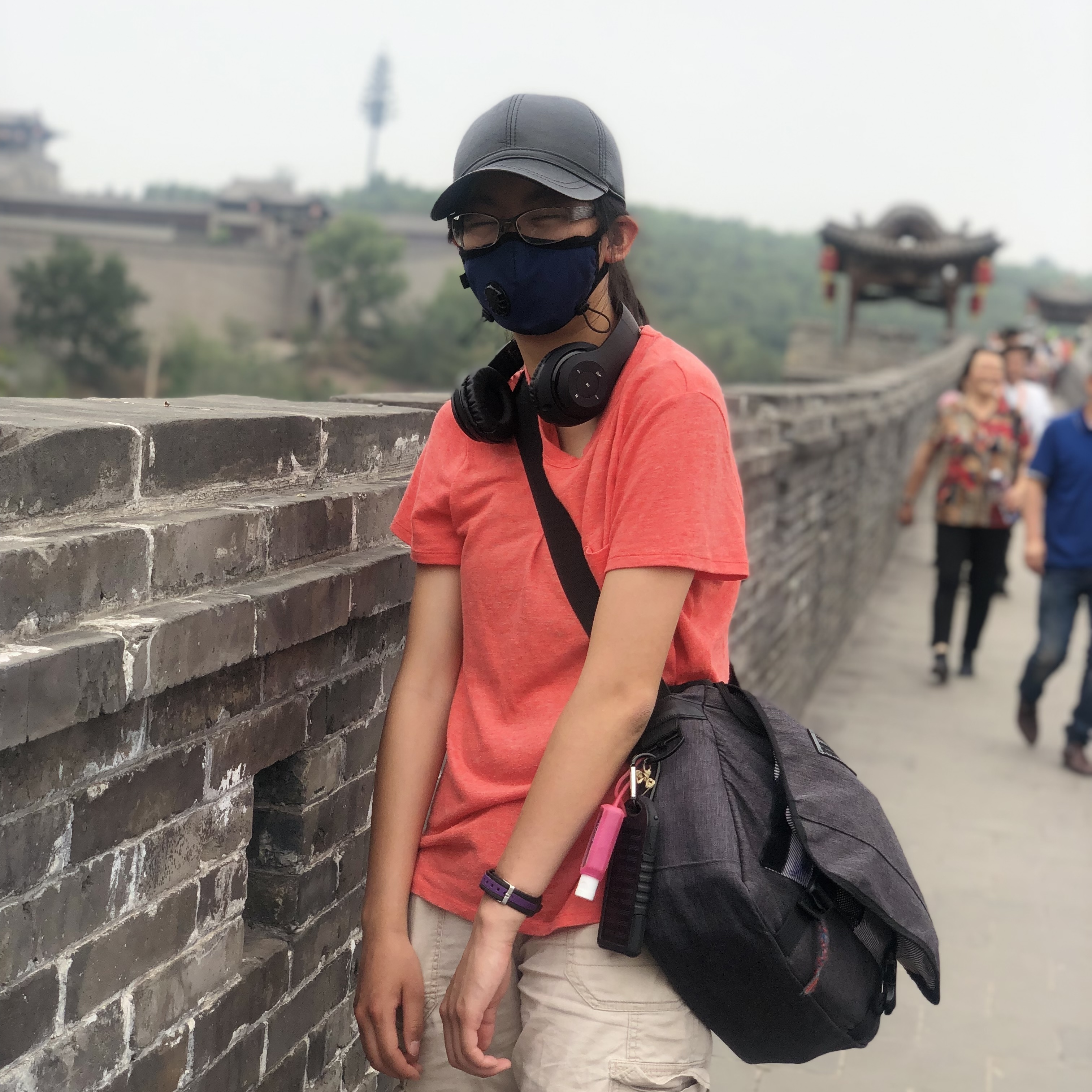 KT Norton stands in front of a stone wall with a shoulder-style backpack, headphones around her neck, a black mask on her face, and a black hat on her head.