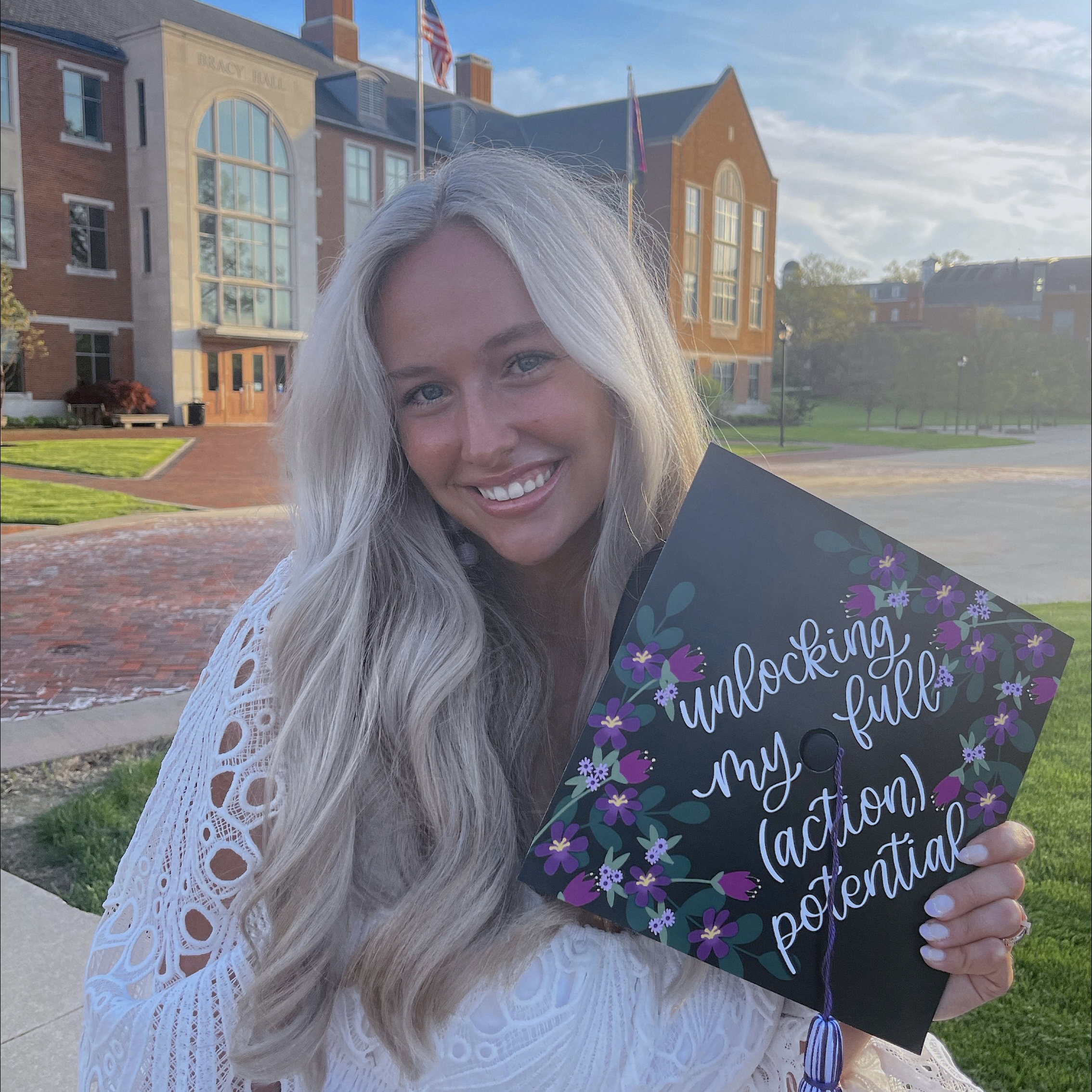 Katie Wilson stands in front of Bracy Hall with a decorated graduation cap with purple flowers. The hat says in white calligraphy-style writing, “unlocking my full (action) potential”